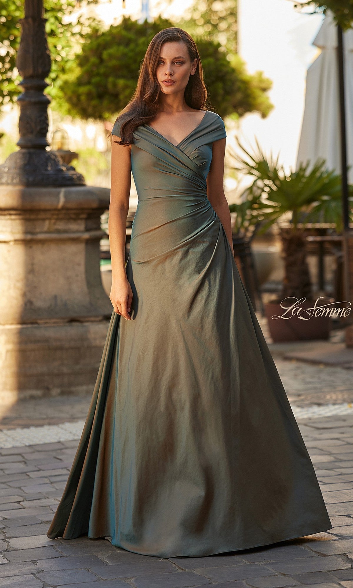 A woman stands outdoors before stone architecture and greenery, wearing the La Femme 33446 long formal dress in moss green—a floor-length taffeta gown with off-the-shoulder draping.